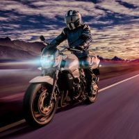 Un motociclista con equipo de protección viaja a gran velocidad por una autopista durante un atardecer vibrante, con montañas de fondo y nubes dramáticas en el cielo.