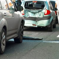 Una colisión trasera que involucra a tres automóviles en una carretera concurrida, con daños visibles en un vehículo. Los transeúntes están parados cerca.