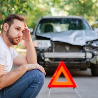 Retrato de conductor caucásico angustiado sentado en el pavimento cerca de su vehículo destrozado tras un accidente automovilístico