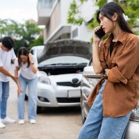 Asian Woman drivers call insurance after a car accident before taking pictures and sending insurance. Car Accident Emergency, while another person tends to a passenger with an injury.