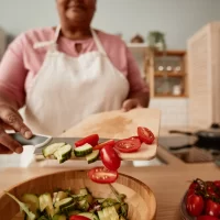 a-person-in-a-pink-shirt-and-white-apron-adds-sliced-cucumbers-and-cherry-tomatoes-from-a-cutting-board-into-a-wooden-bowl-of-salad[1]